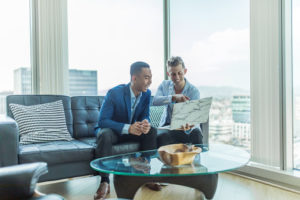 men sitting on couch with laptop, large class windows, coffee table, wooden bowl, blue blazer, marble